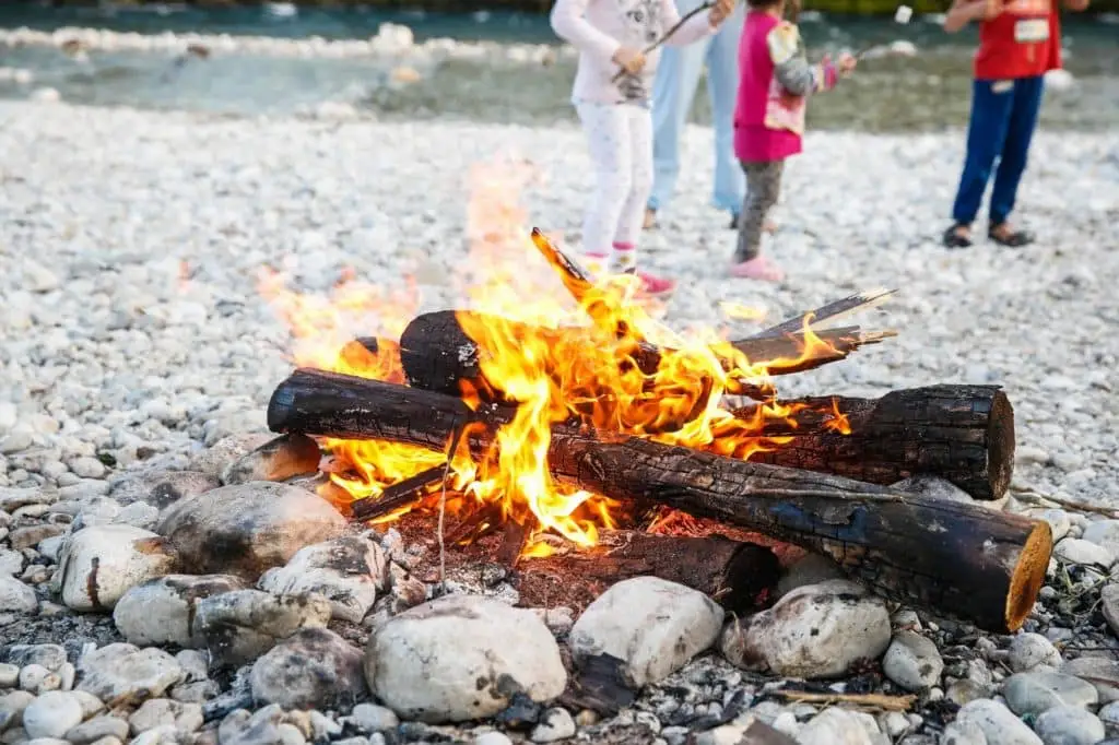 A cozy campfire burns brightly with flames and embers on a rocky shore, as people enjoy the warmth and ambiance in the background.