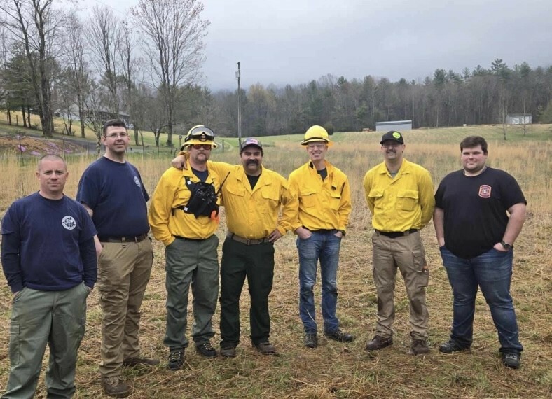 A group of seven men, some wearing uniforms and yellow shirts, stand together in a grassy field. The background features trees and a cloudy sky. The men are smiling, and some are wearing hats and sunglasses.