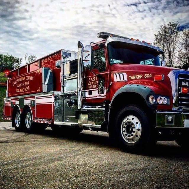 Red fire tanker truck labeled "East Sullivan County Fire Dept" parked on a road. The truck is shiny and well-polished, with trees in the background and a cloudy sky above.