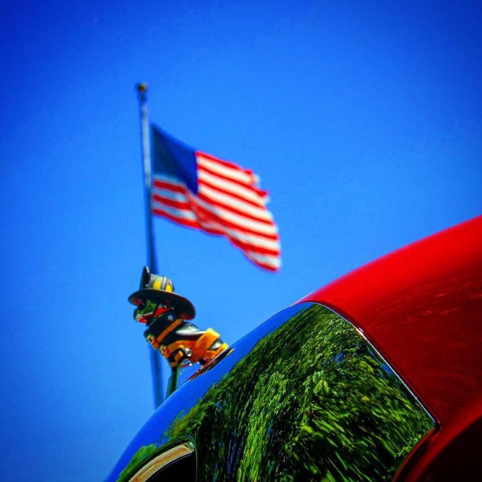 Close-up of a shiny red car hood with a firefighter-themed hood ornament. In the background, an American flag waves against a clear blue sky. The ornament reflects greenery from the surrounding area.