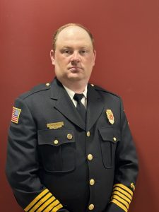 A man in a formal dark navy uniform stands against a red wall. The uniform features a badge, gold buttons, an American flag patch, and gold stripes on the sleeve, indicating a high-ranking fire department official.