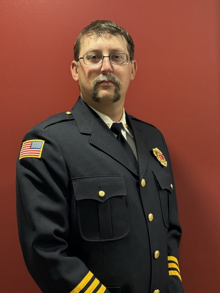 A man with glasses and a mustache wearing a formal dark uniform with gold buttons, a badge, and an American flag patch on the sleeve stands against a solid red background.