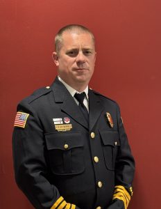 A man in a formal dark uniform with badges, an American flag patch, and gold stripes on the sleeves stands against a plain red background, looking at the camera with a neutral expression.