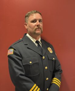 A man in a dark formal uniform with gold buttons and badges stands against a plain red background. He has short hair, a mustache, and an American flag patch on his sleeve.