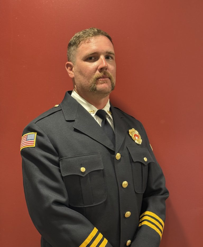 A man in a dark formal uniform with gold buttons and badges stands against a plain red background. He has short hair, a mustache, and an American flag patch on his sleeve.