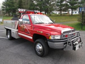 A red fire department utility vehicle with a front-mounted grill guard and emergency lights, parked on a paved area. It has metal storage compartments on the sides and trees in the background.
