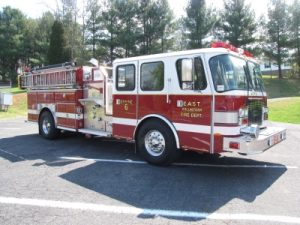 A red and white fire truck parked on a road with trees in the background. The truck is labeled "East Volunteer Fire Dept. Engine 6" and has various compartments and equipment visible.