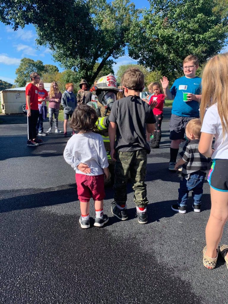 A group of children and a firefighter engaging together outdoors on a sunny day. The kids are facing the firefighter, who is wearing full gear, while trees and other people are visible in the background.