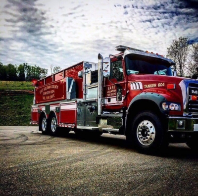 A red and white fire truck labeled "TANKER 604" is parked on a paved surface under a partly cloudy sky. It has multiple wheels and a large tanker section. Trees and grassy hills are in the background.