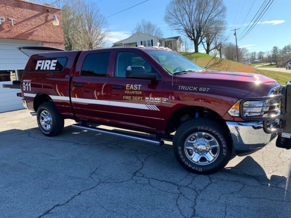 A red volunteer fire department truck is parked on a paved area with "East Volunteer Fire Dept" and "Truck 607" on the side. It has a 911 emergency number and sits in front of a building and a grassy hill under a clear sky.