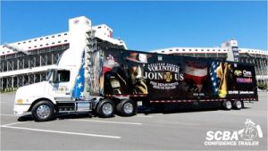A white semi-truck with a large trailer featuring recruitment images and text for a fire department is parked in an empty lot. The building in the background is a multi-level structure with multiple windows. Text on the trailer reads "Join Us.