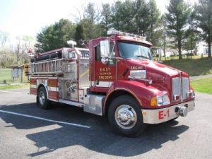 A red fire truck parked on a paved surface, with "East Volunteer Fire Dept" and "Engine 1" labeled on its side. The truck has equipment compartments and is surrounded by trees and a grassy area in the background.
