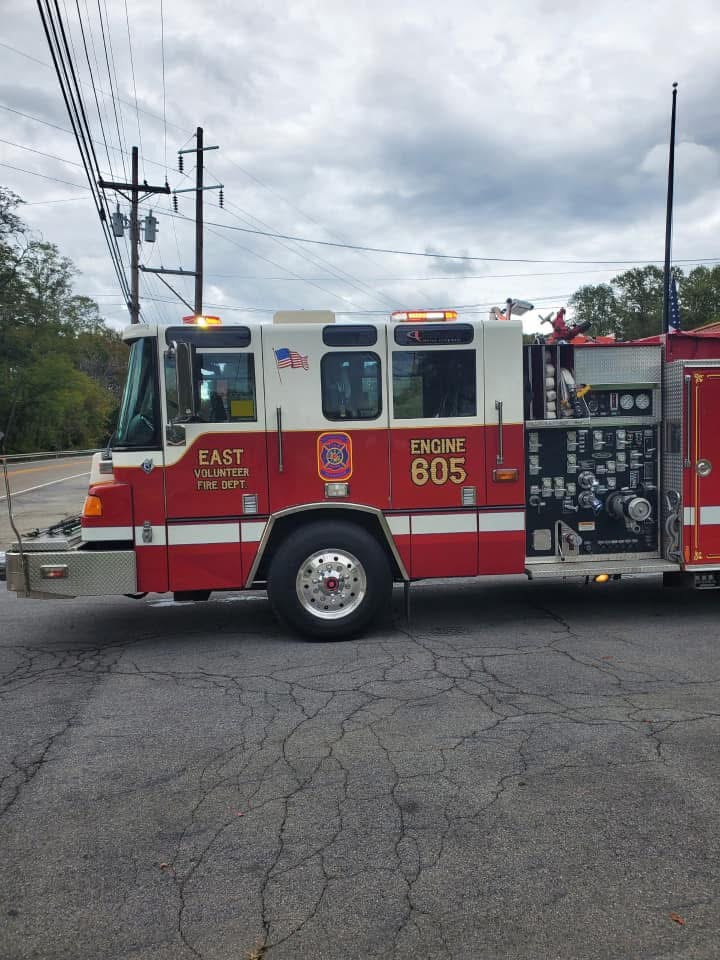 A red and white fire truck labeled "EAST Volunteer Fire Dept. Engine 805" is parked on a cracked asphalt road, with power lines and trees visible in the background under a cloudy sky.