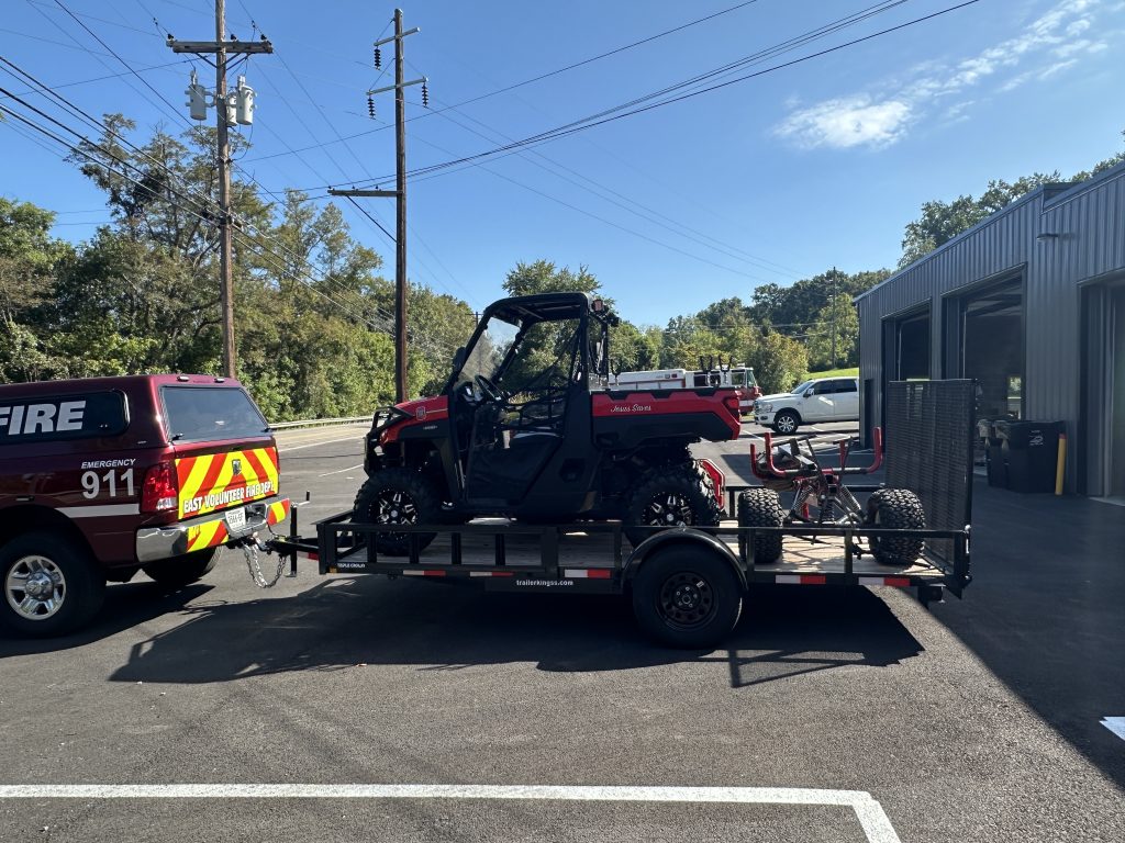 A red emergency vehicle is parked next to a trailer carrying an all-terrain vehicle (ATV) outside a metal building on a sunny day. Trees and utility poles line the road in the background.