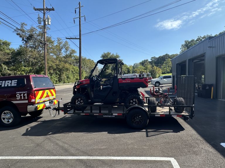 A red emergency vehicle is parked next to a trailer carrying an all-terrain vehicle (ATV) outside a metal building on a sunny day. Trees and utility poles line the road in the background.