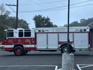 A red and white fire rescue truck labeled "East Fire Dept Rescue 608" is parked on a paved lot with trees and power lines in the background.